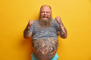 Joyful bearded man with fat belly, clenches fists, happy as won competition, has tattoos, dressed...