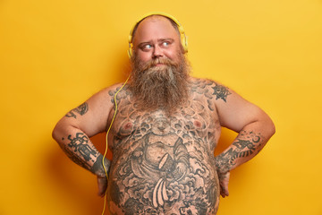 Pensive fatso man keeps hands on hips, listens music for soul in headphones, has tattooed body,...