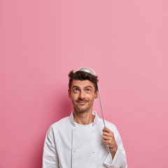 Positive male chef poses with ladle on head, going to prepare soup, wears white uniform, holds kitchen utensils, makes restaurant cuisine, looks above, poses over rosy background, empty space
