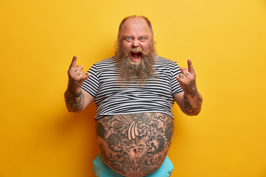Indoor shot of overweight rock fan gestures actively and shouts, enjoys favorite music, keeps mouth widely opened, shows horns hand gesture, has very big tattooed belly, stands against yellow wall