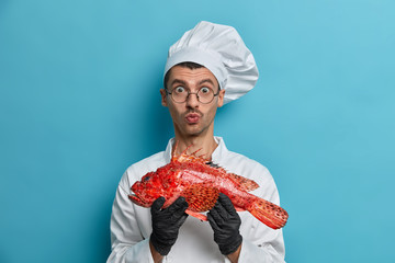 Cooking preparation, culinary concept. Surprised chef holds red sea bass, wears transparent glasses, cook uniform and black gloves, ready to prepare new fish dish, healthy food poses at kitchen indoor
