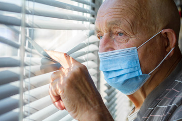 Very elderly man in blue protective mask on his face looks out through the window, stay at home concept