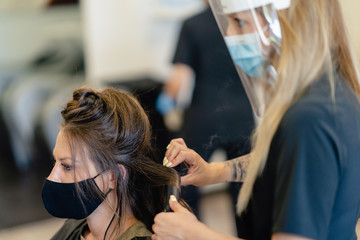 Fototapeta premium Hairdresser, protected by a mask, combing her client's hair with a hair iron in a salon.