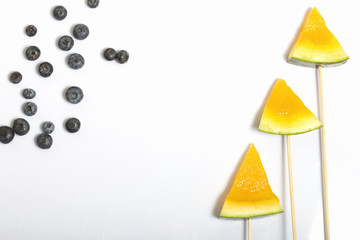 Pieces of yellow watermelon on sticks. Nearby are blueberries. Plate on a white surface.