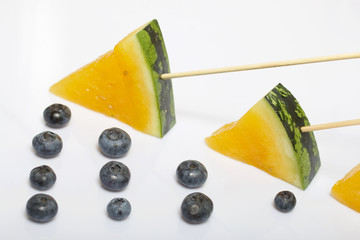 Pieces of yellow watermelon on sticks. Nearby are blueberries. Plate on a white surface.