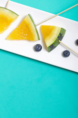 Pieces of yellow watermelon on sticks. Lie on a plate. Nearby are blueberries. Plate on a mint colored surface.