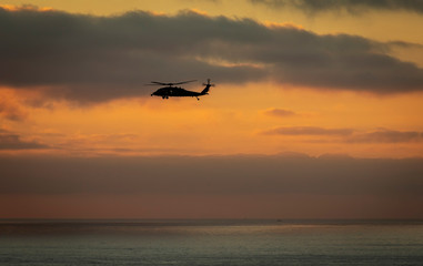 A military helicopter flying toward the left with a cloudy sunset over the pacific ocean