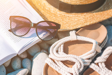 Beach hat on opened book with sunscreen and shoes on pebble beach