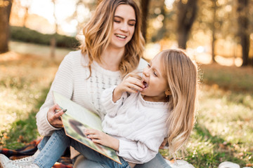 Happy beautiful mother with daughter is having fun and read book sitting in park.