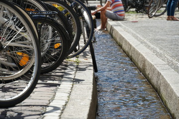 Freiburg old town water channel with bicycles  parked on one bank. A man sitting on the other side is cooling and refreshing his feet of the flowing water of the rill on a hot summer day. 