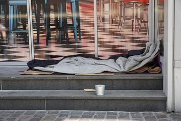 Sleeping bag of a homeless person spread on the stairs in front of a modern posh lounge or restaurant. There is a small metal cup near the berth asking for alms and charity.