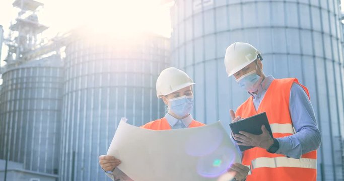 Portrait of male and female engineers in helmets and face masks standing outdoor with graphic and discuss industry project in fabric. Man typing on tablet and woman controller looking at plan