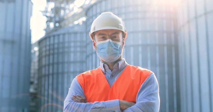 Close up portrait of handsome Caucasian male constructor in helmet and uniform standing in front of big cisterns in factory yard. Man engineer in face mask looking at camera. Construction concept