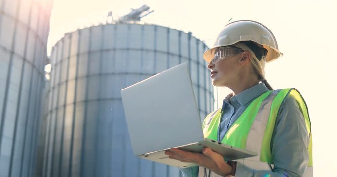 Close up portrait of beautiful joyful female controller working on laptop and looking away. Woman constructor in helmet tapping on computer outdoor near big tanks in plant. Engineer concept
