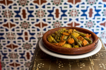 Moroccan vegetable tajine on wooden table, with traditional arabic tiles in the background.