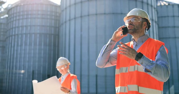 Portrait of Caucasian man engineer in helmet speaking on smartphone. Male constructor calling on cellphone outdoor. Female controller in uniform looking at plan in plant yard. Construction concept