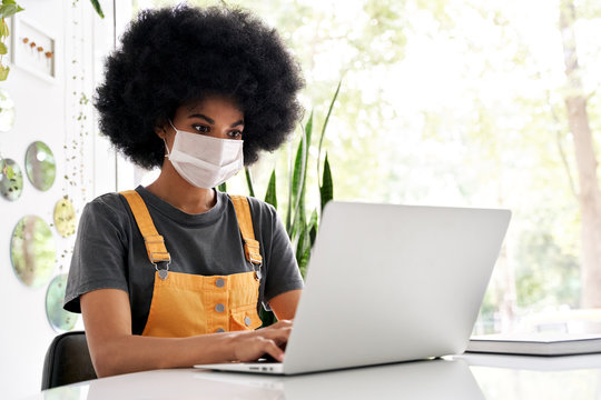 Young African American Teen Girl Student With Afro Hair Wearing Face Mask Using Laptop Sitting At Cozy Cafe Table Alone Indoor. Social Distancing And Work, Study On Computer With Covid 19 Protection.