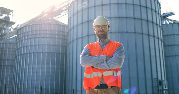 Portrait of handsome Caucasian male constructor in helmet and uniform standing in front of big cisterns in factory territory. Man engineer looking at camera at plant outdoors. Worker concept