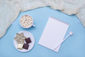 Christmas hot drink with marshmallows and chocolate candies on blue background surrounded by white knitted scarf. Blank sketchbook for notes. Cozy winter concept. Place for text
