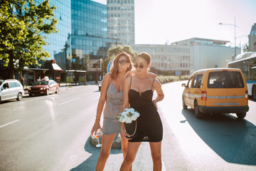 Two attractive caucasian women in summer tight dresses pose with flower boxes and cocktail on the road. Fashion and the city