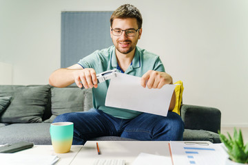 Front view on young adult caucasian man entrepreneur sitting on the couch sofa at home using stapler on paper documents letter or invoices - Male work from home in his room