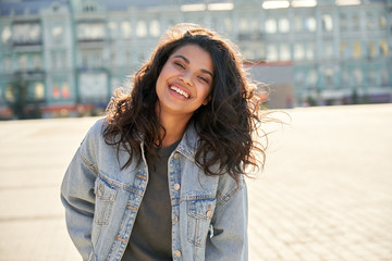 Happy African young woman wearing denim jacket laughing looking at camera standing on street. Smiling Afro American teen generation z hipster girl posing outdoor backlit with sunlight, portrait. - obrazy, fototapety, plakaty