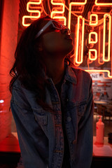 Hipster cool African American teen gen z girl wears trendy glasses posing under neon night sign...