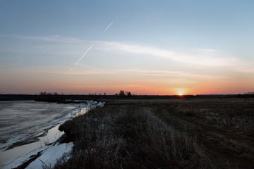 Landscape. Early spring morning, sunrise. Gray ice on the river, old grass in the field, white airplane footprints in the blue sky.