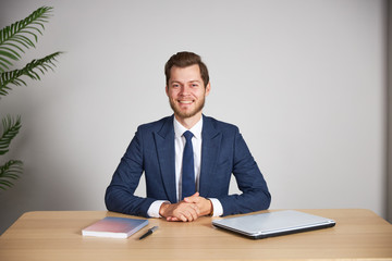 The businessman sits at his desk on a white blank background, crosses his arms and looks into the camera, smiling.