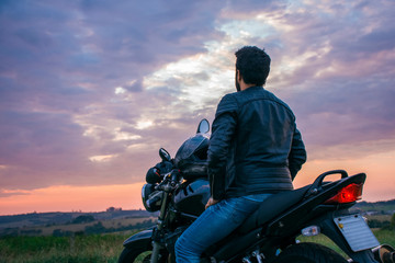 Man sitting on a black motorcycle, wearing blue jeans, a black jacket and a black helmet, with his back turned against a landscape in the background.