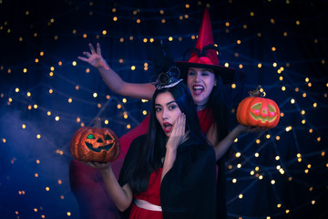 o Women Dressed as Witches Holding Jack-o'-Lanterns and Lantern, Celebrating Halloween with Spooky Costumes and Festive Lights in the Background
