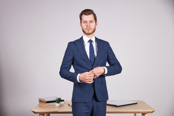 A businessman in a blue business suit stands in front of a wooden office table and clasps his jacket. White background.