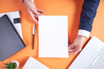 Orange office desktop with business accessories. Laptop, books, pen. The businessman is holding a white sheet of paper with an empty background. Flatley is a business of details.