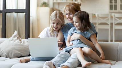 Fototapeta na wymiar Happy together. Mother or millennial female babysitter sitting on sofa hugging two little laughing sisters watching fun movie on laptop, playing education game, studying language course for children