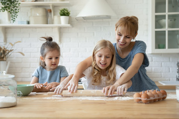 Happy young mother showing elder daughter how to roll dough at kitchen table and younger girl funny clumsy little kid trying to repeat after sister, millennial babysitter teaching children to bake