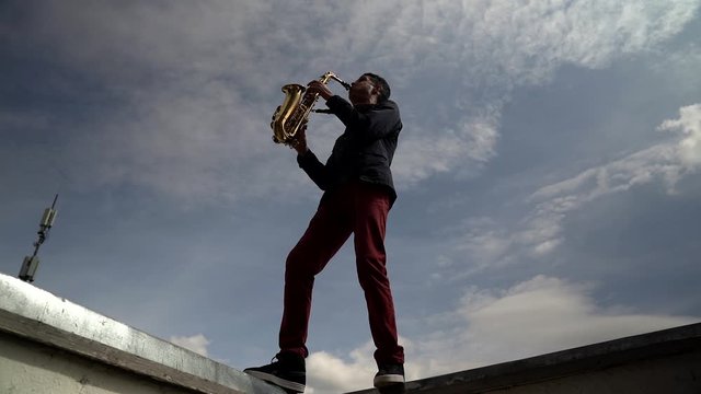 The silhouette of a young stylish guy playing a saxophone on the roof of a high-rise building against the sky. Wide view from below