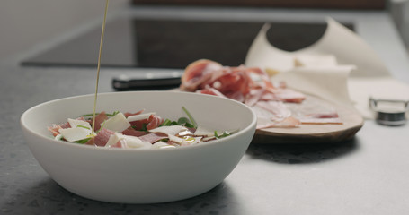 pour olive oil over salad with prosciutto, cherry tomatoes and mixed salad leaves in white bowl