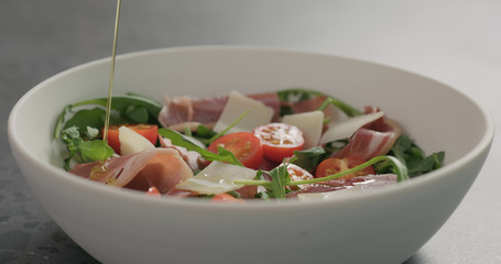 pour olive oil over salad with prosciutto, cherry tomatoes and mixed salad leaves in white bowl