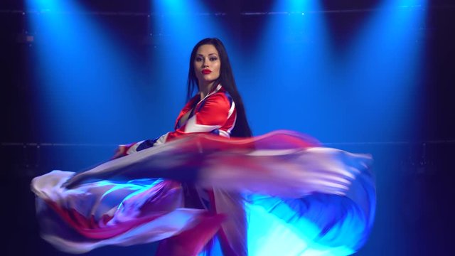 Passionate girl dancing in a sexy suit in the color of the English flag and rhinestones. Attractive brunette waving her skirt in a dark studio with smoke and blue neon lights. Female dance show.