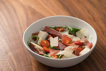 Salad with prosciutto, tomato, arugula leaves and parmesan cheese in white bowl on walnut table