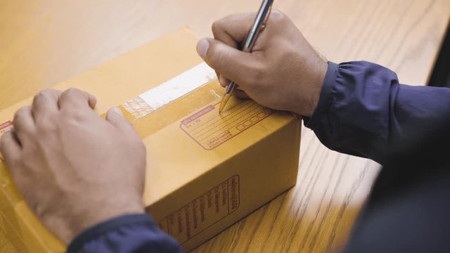 Close Up. Fast post shipment service concept. young asian man customer writing recipient address on parcel cardboard box at home office desk. 4K Resolution.
