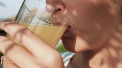 Woman is drinking orange juice. Side view of female hand bring glass with orange juice to their mouth and drink it on sunny day in nature. Close-up