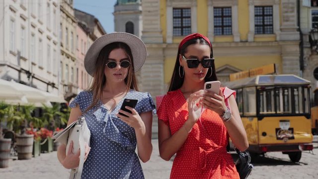 Two cheerful stylish and friendly girls in sunglasses walking the city street and using smartphone for likes, followers, comments for social media.