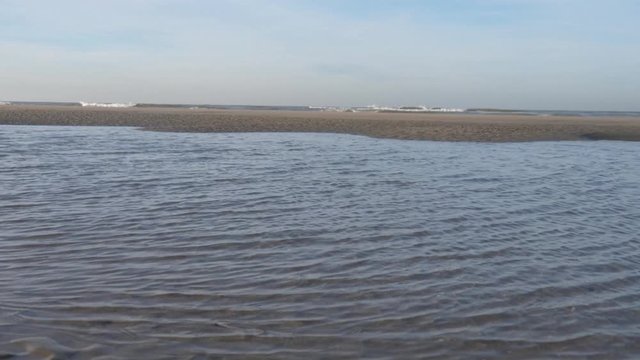 Remnants of water driven by the wind with small waves, view of a sandbank and the surf, behind it the open North Sea and the horizon, a single bird briefly flies through the picture