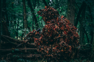 Fully covered sniper in a green forrest. Hiding behind a log. Air-soft, military, warfare. 