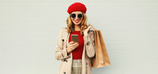 Autumn portrait of smiling woman with smartphone and shopping bag wearing a coat, red french beret over gray background