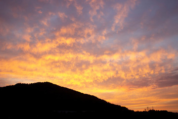 A fiery orange-red sunset. Ragged Cirrus clouds in the dark blue night sky. Black silhouette of a mountain. Amazing phenomenon. Background.