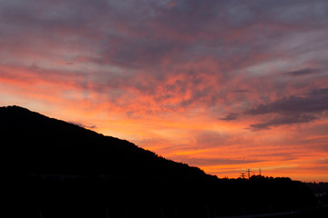 A fiery orange-red sunset. Ragged Cirrus clouds in the dark blue night sky. Black silhouette of a mountain. Amazing phenomenon. Background.