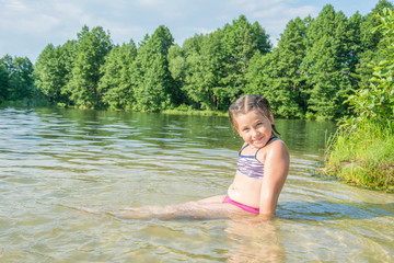 In the summer on the lake during the day, a little girl with pigtails sits in the water.