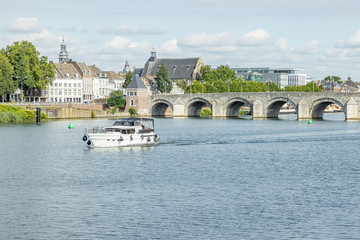 Naklejka premium Maas river with a boat sailing, the Servatius bridge (Sint Servaasbrug) and with an urban landscape of the city of Maastricht in the background, sunny day in the South Limburg, Netherlands